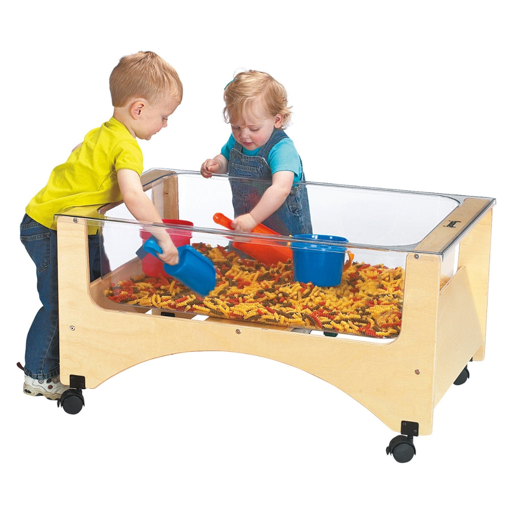 Two children playing with a transparent sandbox table filled with dried pasta and toys on a white background.