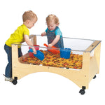 Two children playing with a transparent sandbox table filled with dried pasta and toys on a white background.